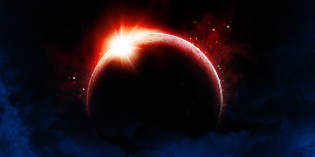 Solar eclipse image with a darkened moon centered in frame, rimmed by a vivid red glow. A bright burst of sunlight breaks over the upper edge, creating a sharp flare, while red particles and nebula-like clouds drift across a deep blue and black space background.